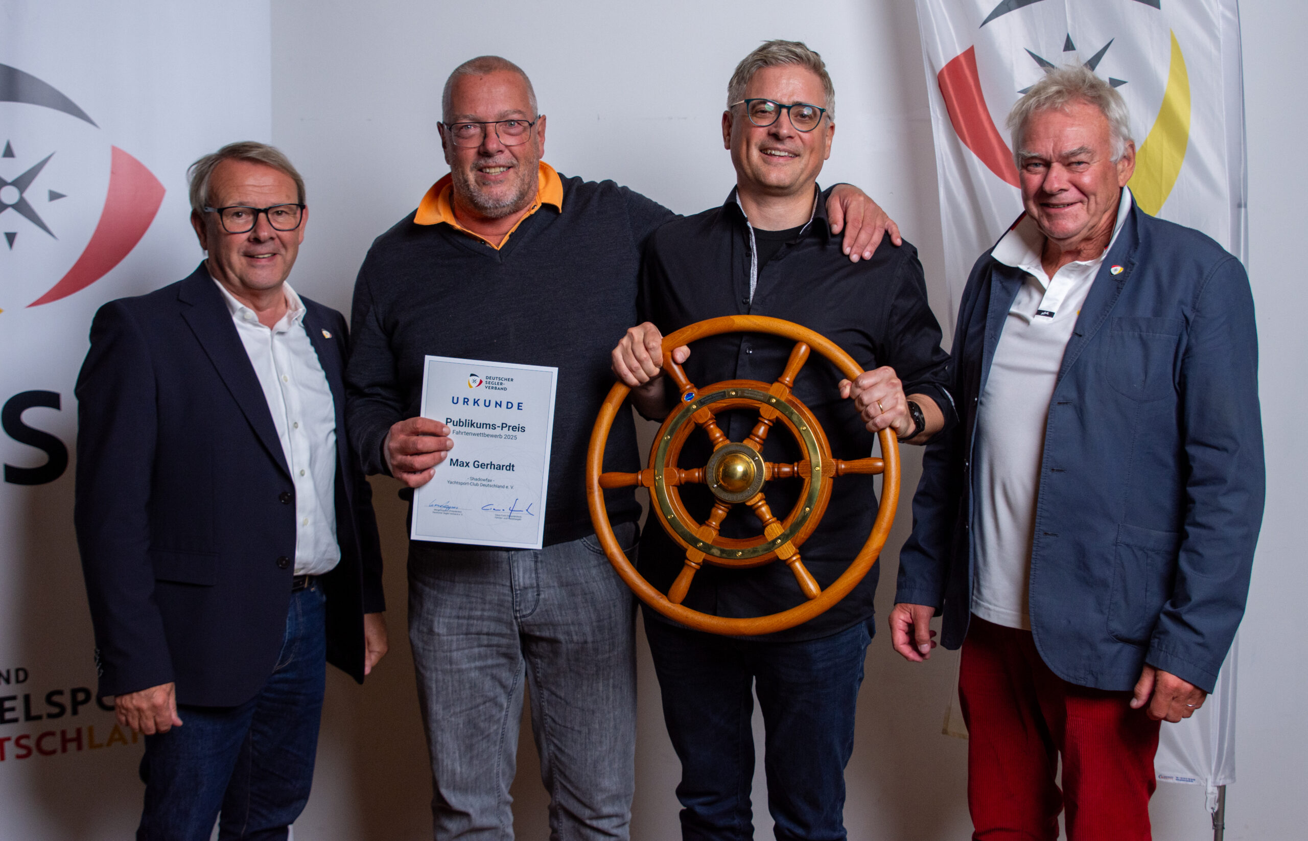 Vier Männer, die beiden mittleren haben den Publikumspreis des DSV gewonnen und halten ein hölzernes Steuerrad in der Hand.