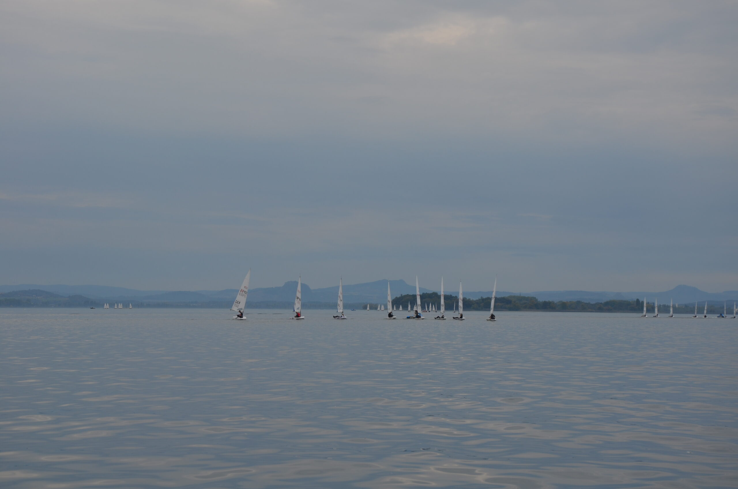 Der Bodensee mit wenig Wind und diesigem Wetter, im Hintergrund die Alpenkulisse