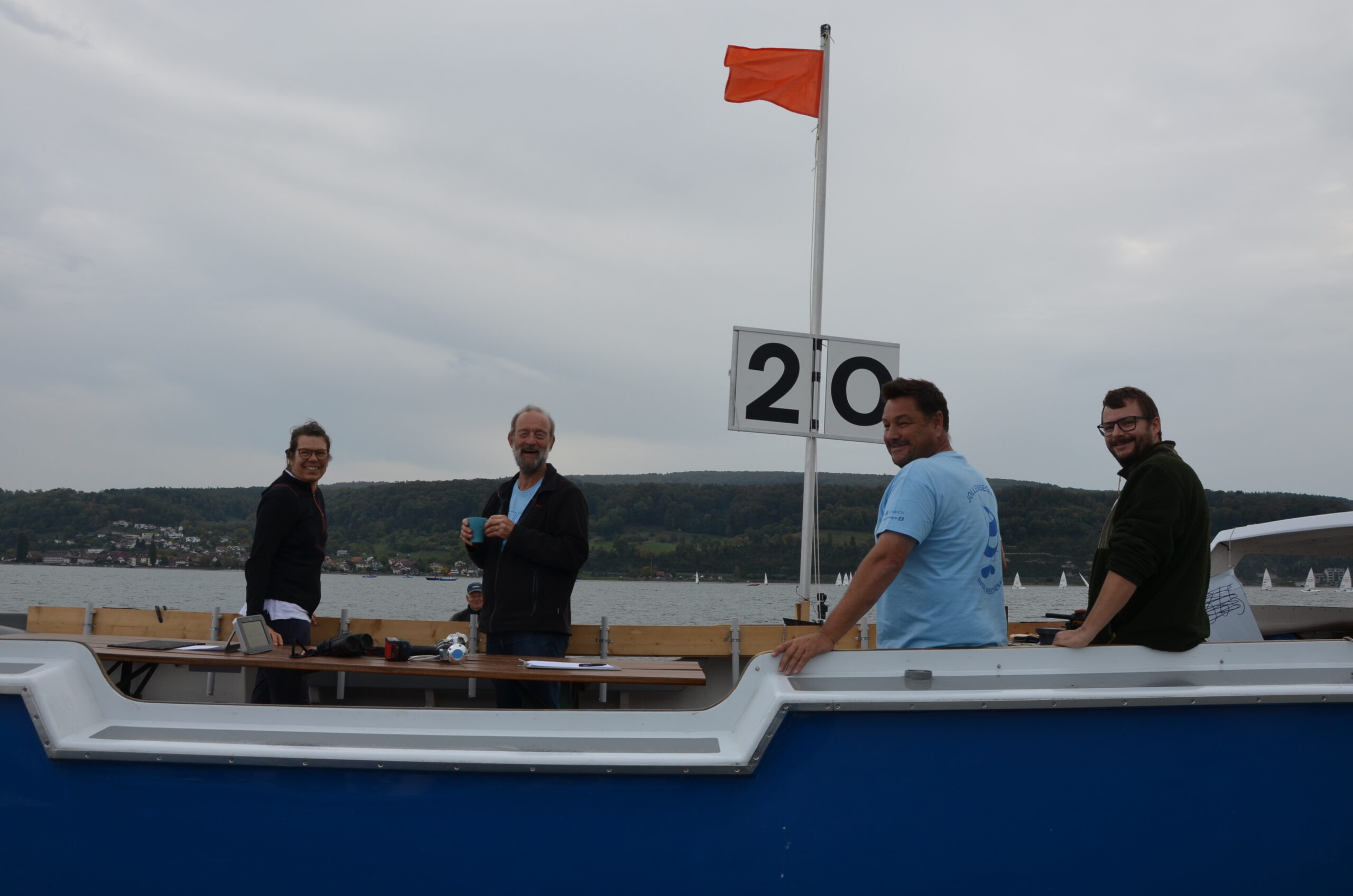 Das Team einer Wettfahrtleitung auf einem blauen Boot, darüber weht eine orangefarbene Flagge.
